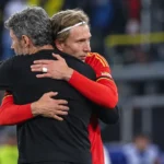 Een Belgische speler in rood omhelst een coach in donkere kleding langs het veld.