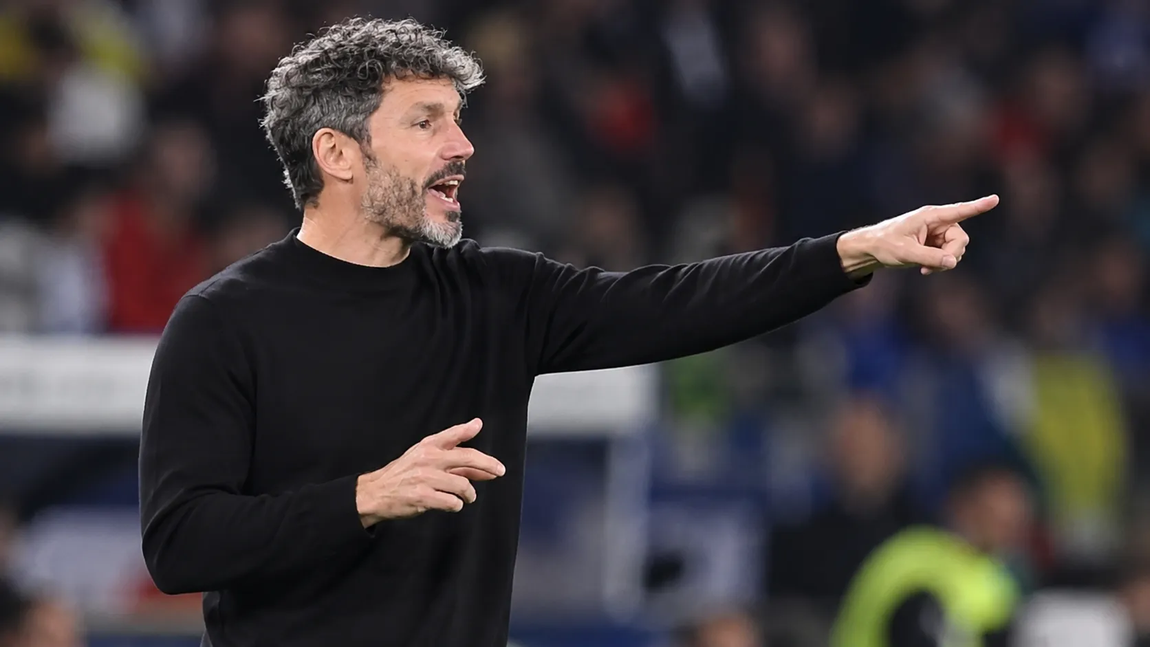 Een coach in zwarte kleding geeft aanwijzingen langs de zijlijn van een interlandstadion.
