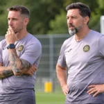Twee leden van een technische staf in trainingskleding langs een oefenveld.
