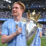 Een speler in lichtblauw tenue houdt een grote trofee vast in een gevuld stadion.