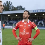 Een speler in rood van KSK Heist met een tegenstander en scheidsrechter op het veld.