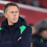Een coach in donkere sportkleding staat in de technische zone van een stadion.