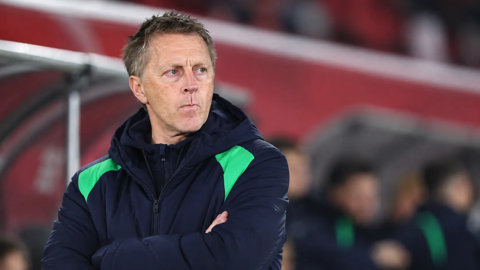 Een coach in donkere sportkleding staat in de technische zone van een stadion.
