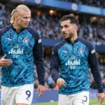 Twee voetballers in blauwe opwarmkleding op het veld van een stadion.