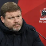 Hein Vanhaezebrouck in een donkere jas in een persruimte met een discreet Jupiler Pro League-embleem op de achterwand.