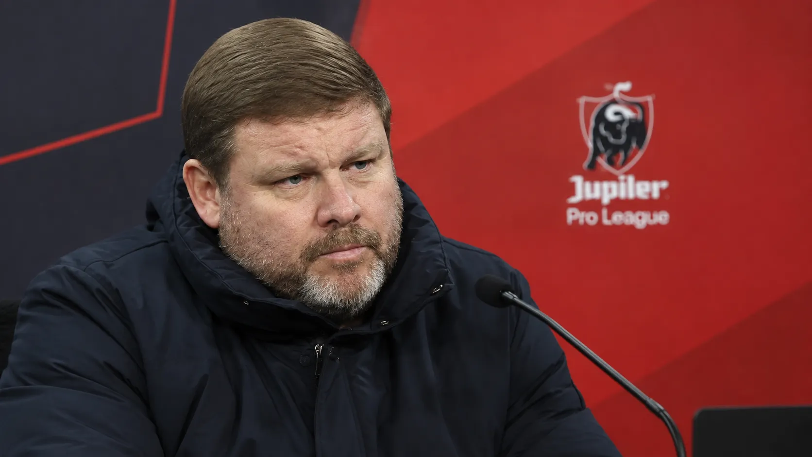Hein Vanhaezebrouck in een donkere jas in een persruimte met een discreet Jupiler Pro League-embleem op de achterwand.