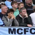 Een groep Manchester City-supporters op de tribune achter een lichtblauwe MCFC-banner.