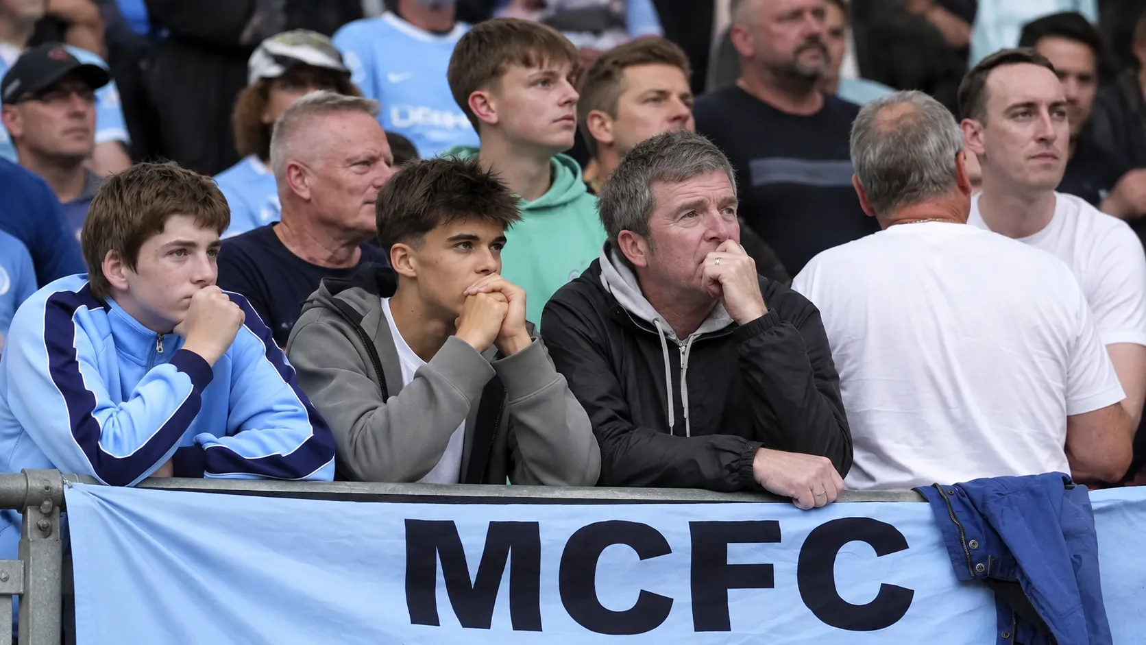Een groep Manchester City-supporters op de tribune achter een lichtblauwe MCFC-banner.