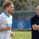 Een Napoli-speler in wit shirt warmt op terwijl een trainer in donker poloshirt toekijkt.
