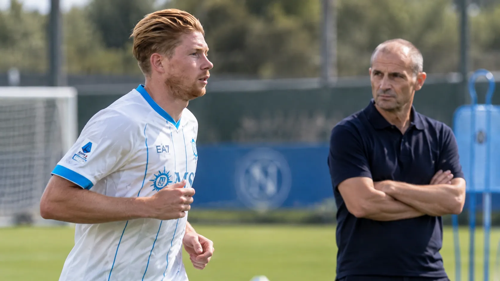Een Napoli-speler in wit shirt warmt op terwijl een trainer in donker poloshirt toekijkt.