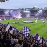 Collectieve Anderlecht-supporters met paars-witte vlaggen in de tribunes.