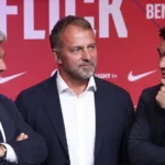 Drie niet-spelende clubvertegenwoordigers in donkere pakken voor een rood persdecor.