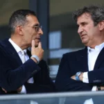 Twee clubofficials in donkere pakken overleggen in een stadionloge.