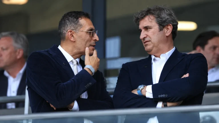 Twee clubofficials in donkere pakken overleggen in een stadionloge.
