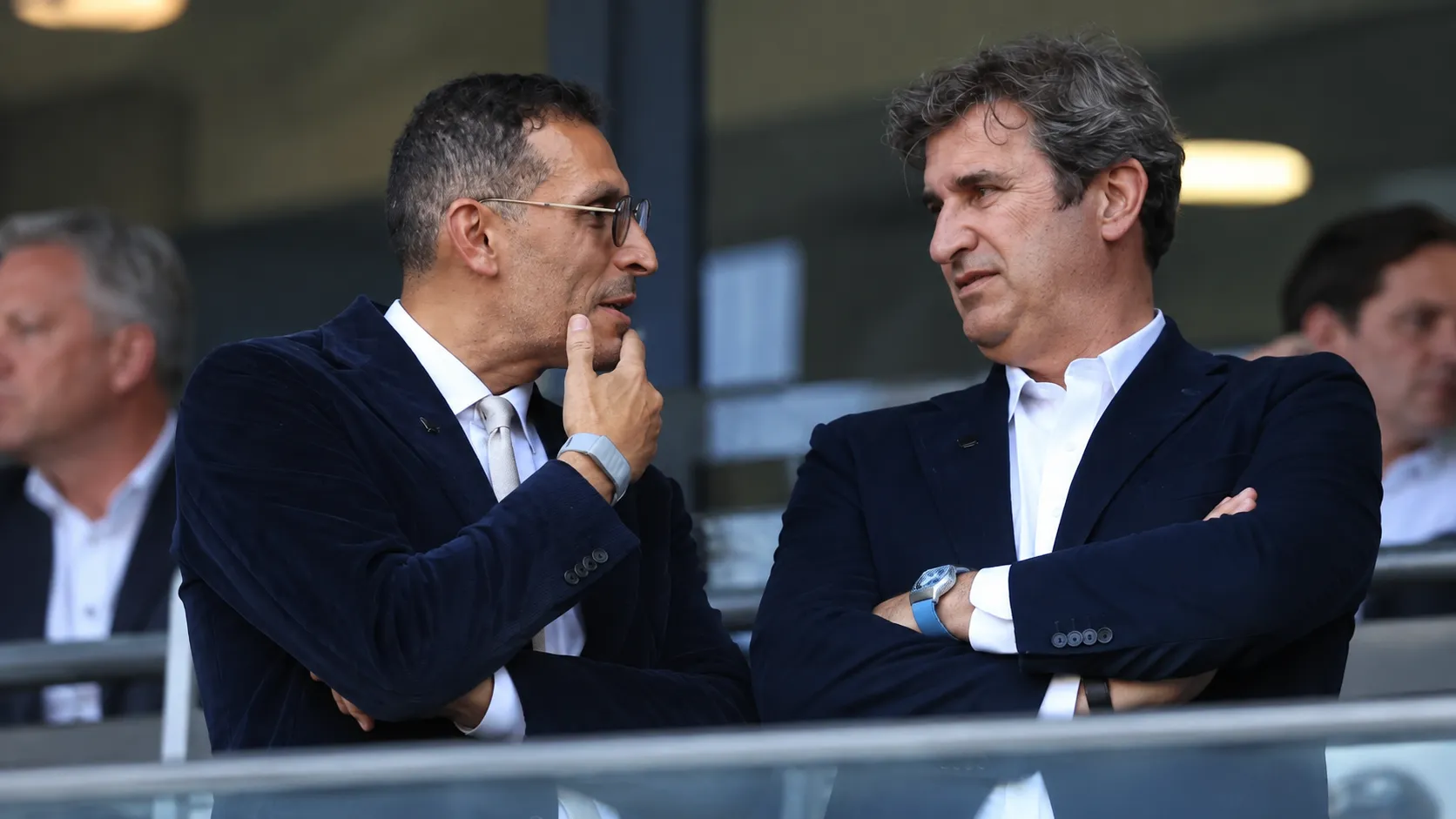 Twee clubofficials in donkere pakken overleggen in een stadionloge.