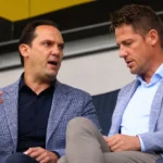 Twee clubofficials in nette kledij in gesprek op de tribune.