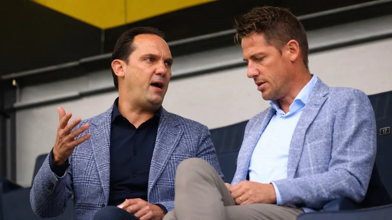Twee clubofficials in nette kledij in gesprek op de tribune.