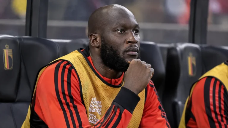 Romelu Lukaku zit in Belgische warming-upkleding op de spelersbank in een stadion.