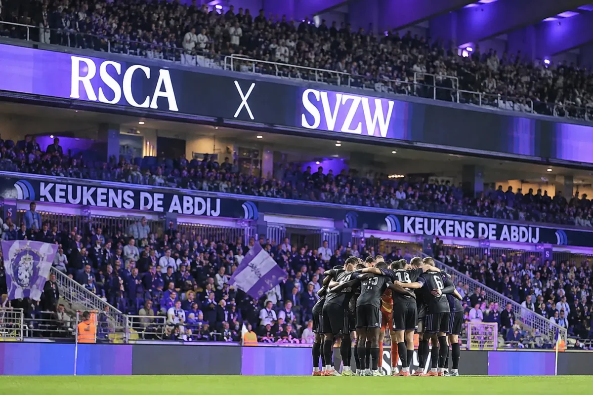Supporters van RSC Anderlecht achter een groep spelers die samenkomt aan de rand van het veld.
