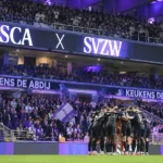 Supporters van RSC Anderlecht achter een groep spelers die samenkomt aan de rand van het veld.