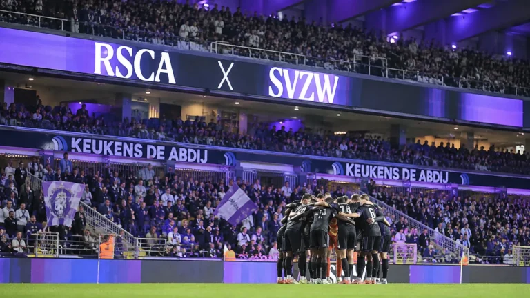 Supporters van RSC Anderlecht achter een groep spelers die samenkomt aan de rand van het veld.