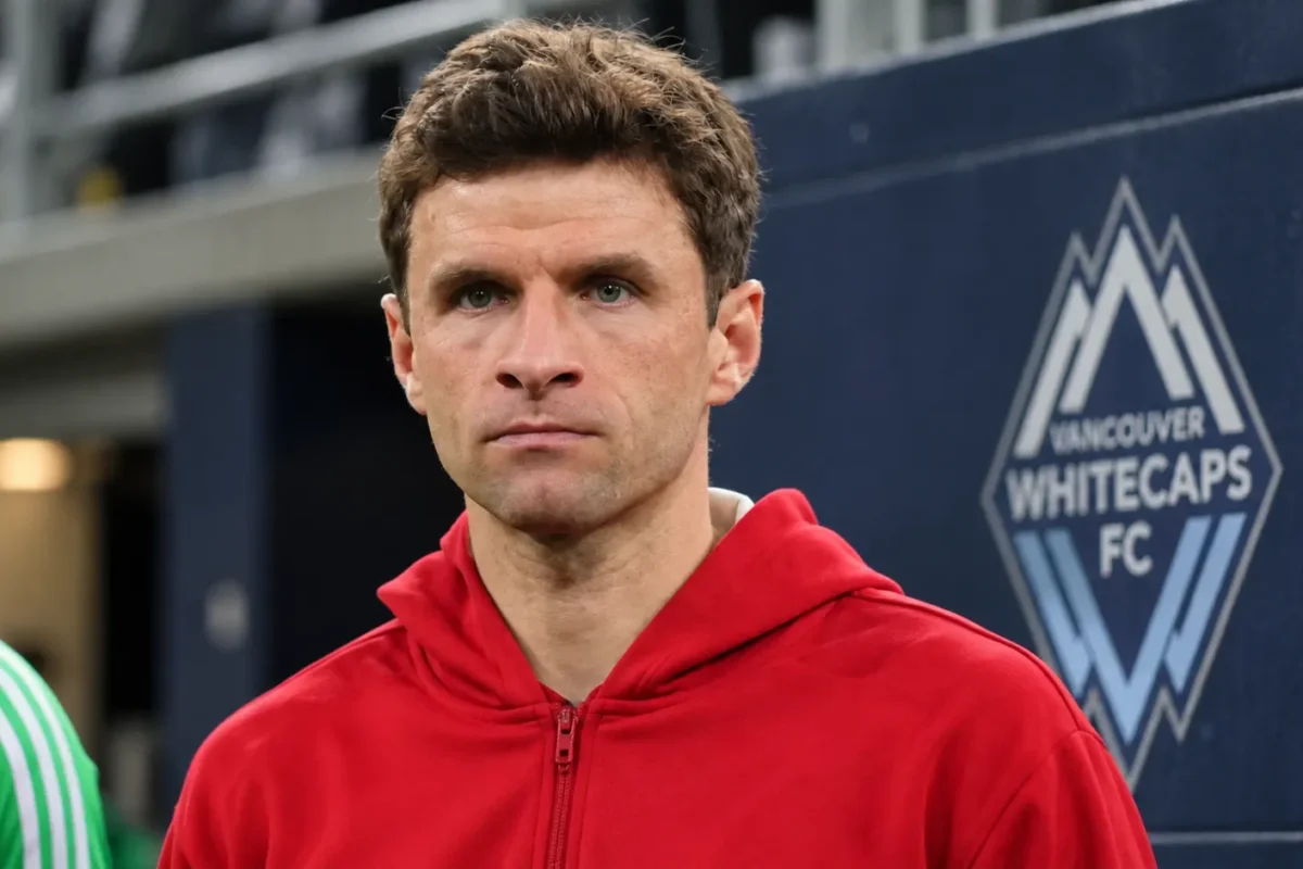 Thomas Müller in een rode sportjas voor een subtiel Vancouver Whitecaps-embleem aan een stadionwand.