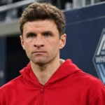 Thomas Müller in een rode sportjas voor een subtiel Vancouver Whitecaps-embleem aan een stadionwand.
