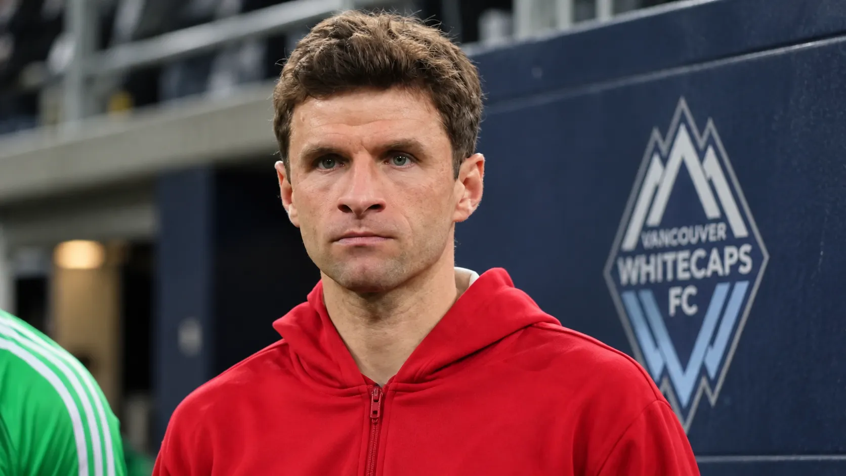 Thomas Müller in een rode sportjas voor een subtiel Vancouver Whitecaps-embleem aan een stadionwand.