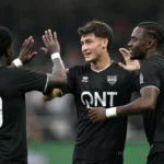 Drie voetballers in zwarte wedstrijdshirts vieren samen op het veld.