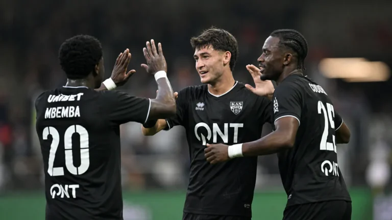 Drie voetballers in zwarte wedstrijdshirts vieren samen op het veld.