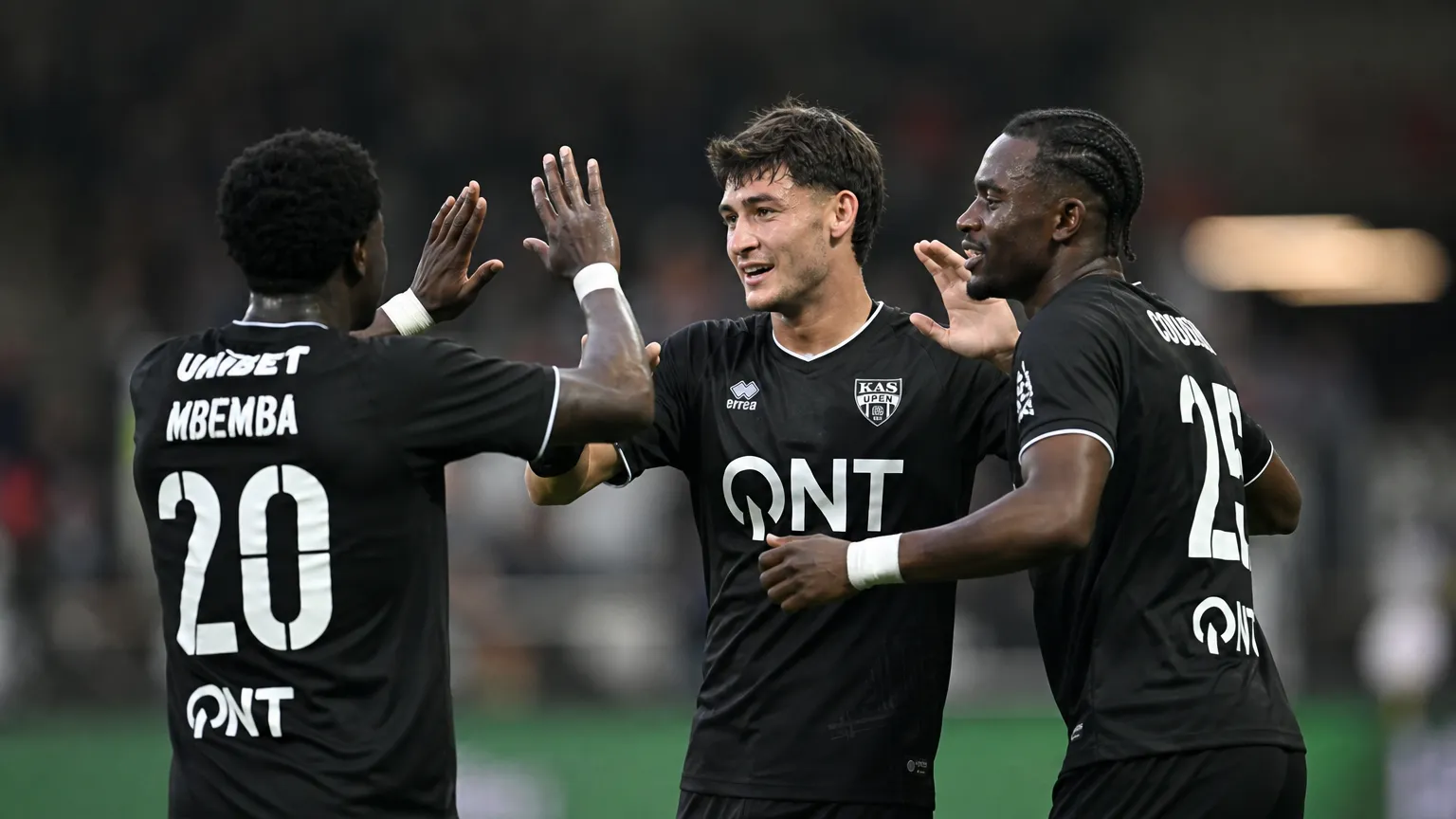Drie voetballers in zwarte wedstrijdshirts vieren samen op het veld.