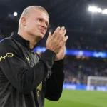 Erling Haaland applaudisseert aan de rand van het veld in donkere Manchester City-kleding.