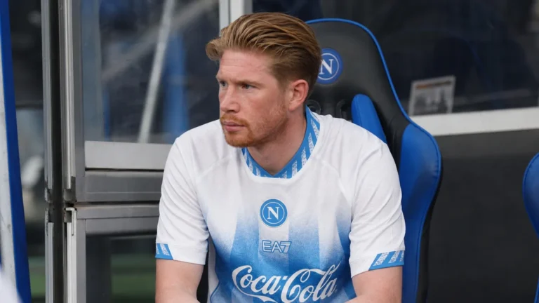 Kevin De Bruyne zit aandachtig op de bank langs het veld in zijn Napoli-wedstrijdshirt.