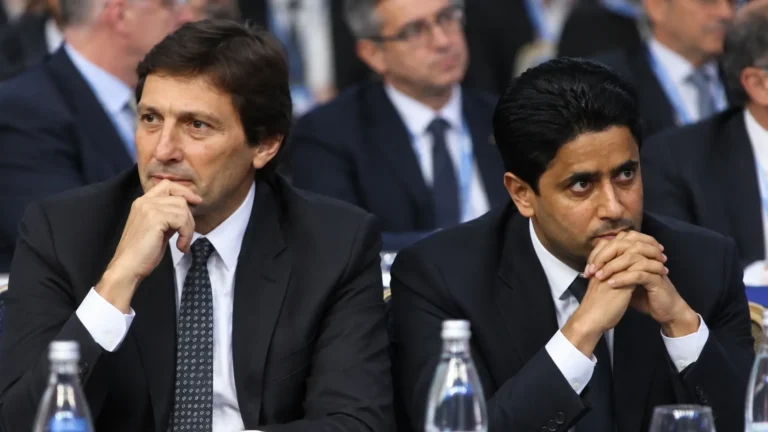 Twee voetbalbestuurders in donkere pakken zitten tijdens een formele bijeenkomst.