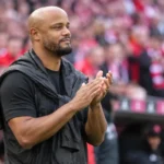 Een coach in zwarte jas applaudisseert langs de zijlijn van een voetbalstadion.