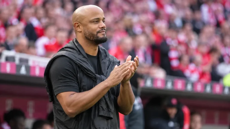 Een coach in zwarte jas applaudisseert langs de zijlijn van een voetbalstadion.