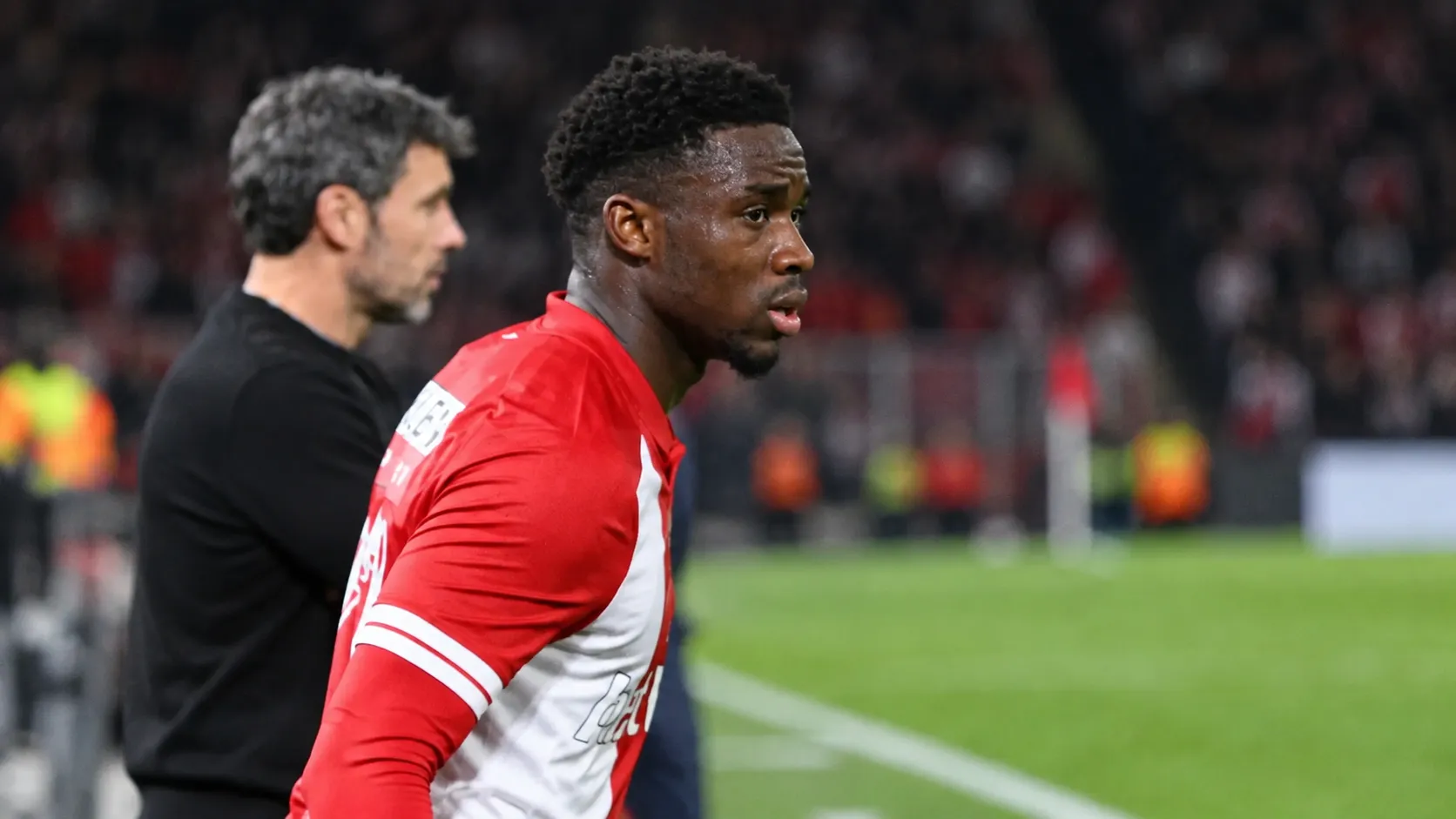 Mandela Keita in een rood-wit wedstrijdshirt langs de zijlijn van een voetbalveld.