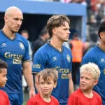 Een groep voetballers in blauwe shirts wandelt met jonge mascottes richting het veld.