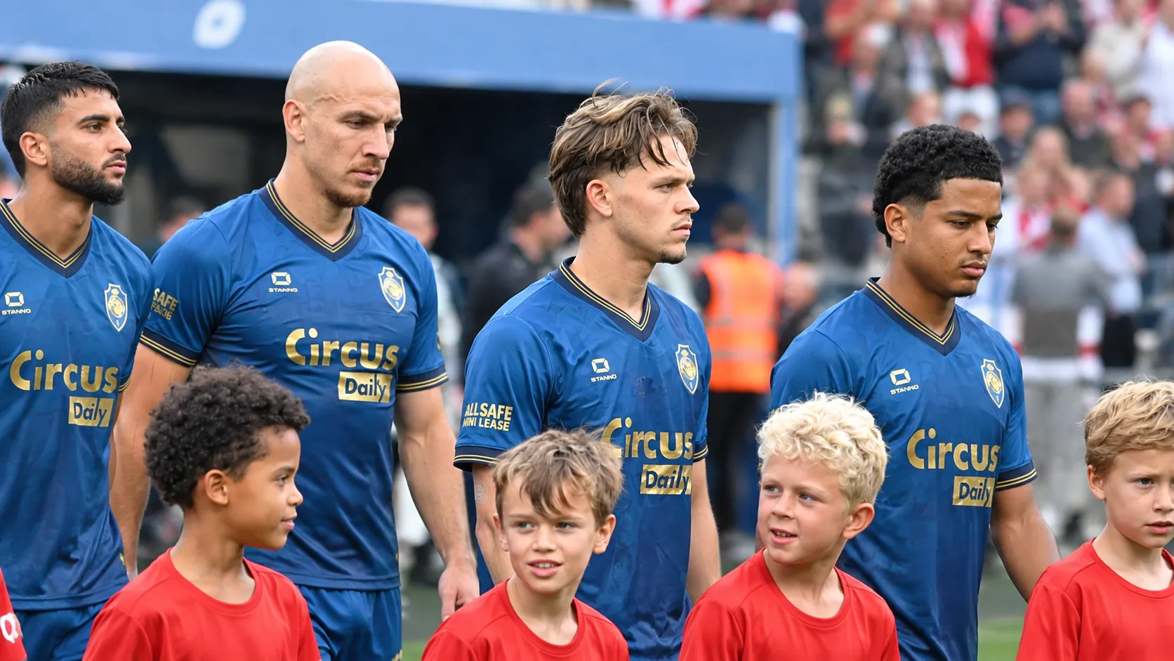 Een groep voetballers in blauwe shirts wandelt met jonge mascottes richting het veld.