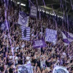 Een volle tribune met paars-witte supporters die applaudisseren.