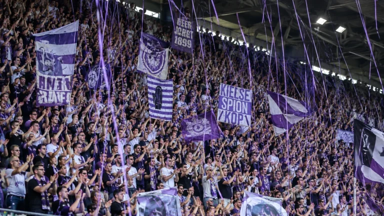 Een volle tribune met paars-witte supporters die applaudisseren.