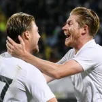 Twee voetballers in witte wedstrijdshirts vieren samen een moment op het veld.