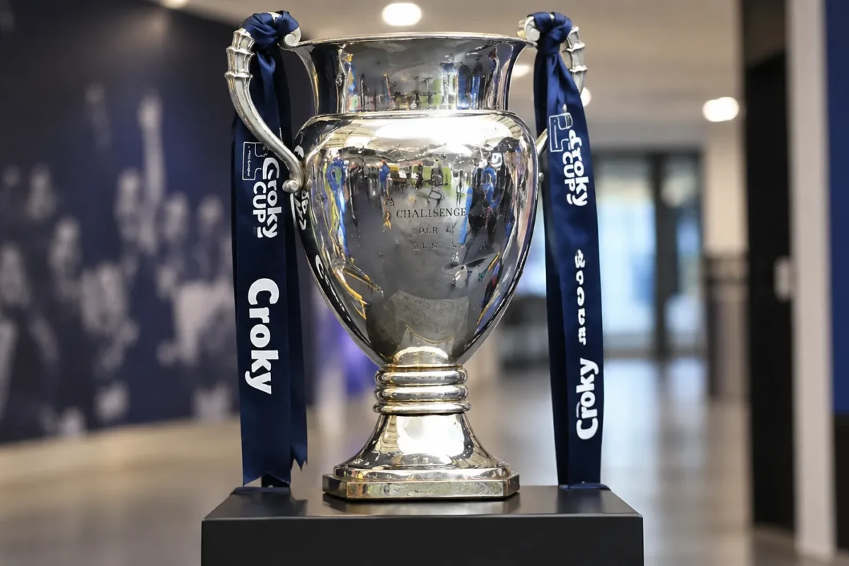 De Croky Cup-trofee met donkerblauwe linten op een sokkel in een indoor voetbalomgeving.