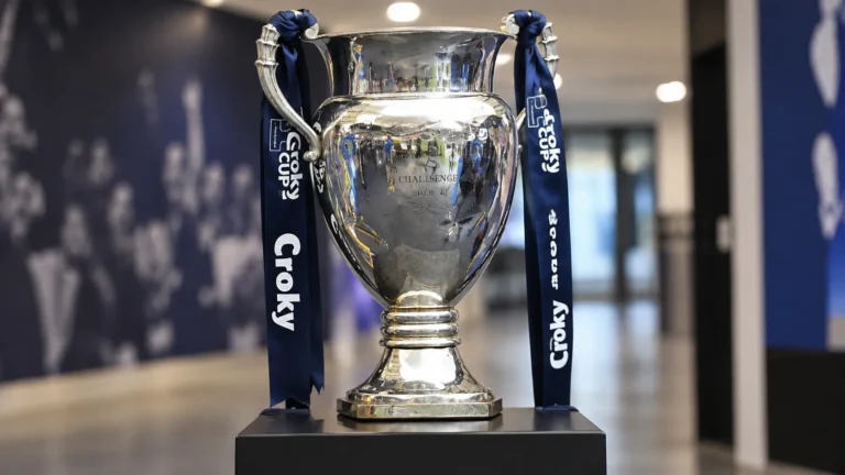 De Croky Cup-trofee met donkerblauwe linten op een sokkel in een indoor voetbalomgeving.