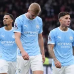 Vier voetballers in lichtblauwe tenues wandelen naast een televisiecamera na een wedstrijd.
