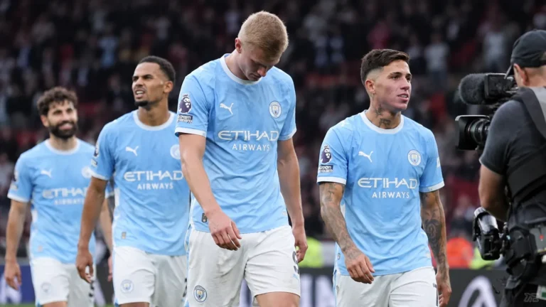 Vier voetballers in lichtblauwe tenues wandelen naast een televisiecamera na een wedstrijd.