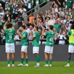 Een groep voetballers in groene shirts applaudisseert voor een volle tribune met supporters.