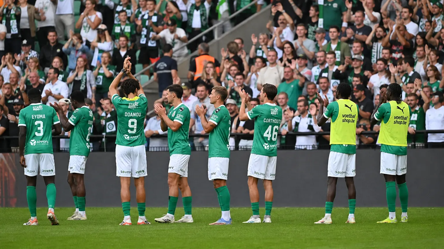 Een groep voetballers in groene shirts applaudisseert voor een volle tribune met supporters.