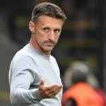 Een coach in een lichtgrijze trui gebaart langs de zijlijn van een voetbalstadion.