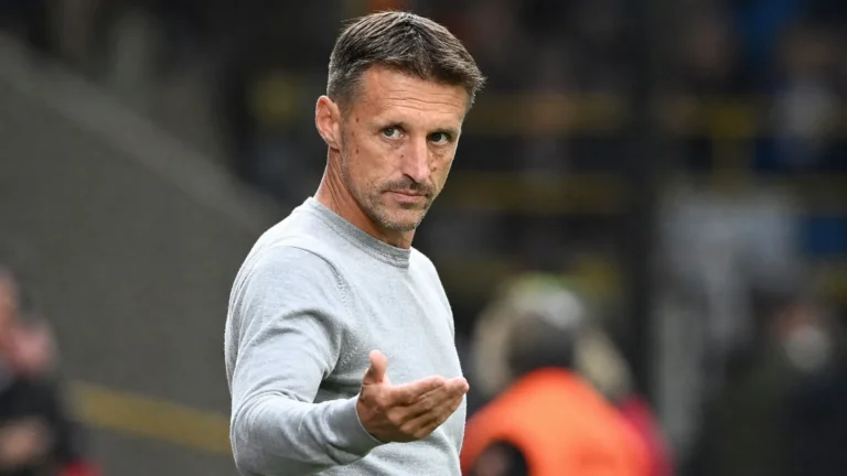Een coach in een lichtgrijze trui gebaart langs de zijlijn van een voetbalstadion.